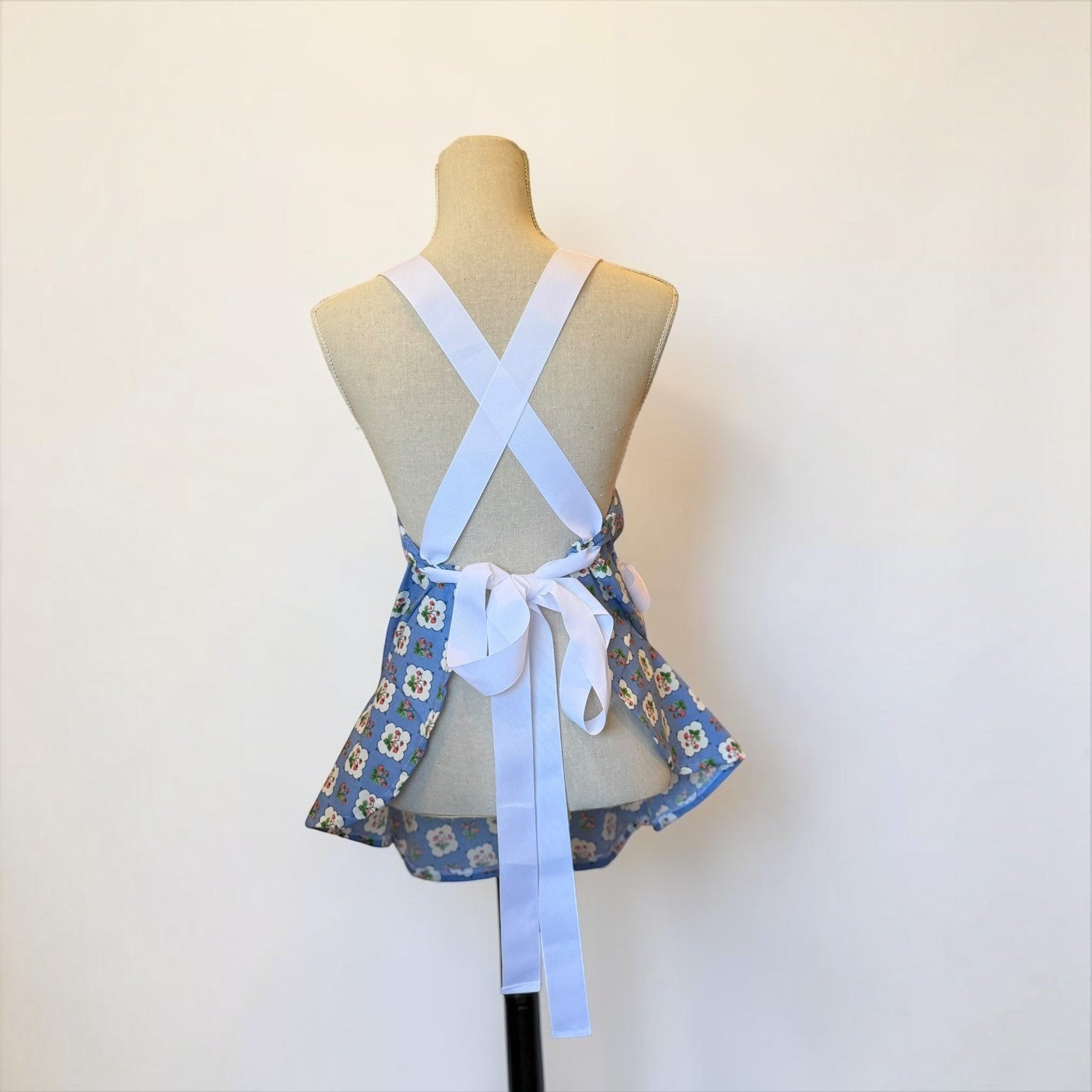 Her Good Eye vintage-inspired blue floral apron with white cross-back straps and a flared peplum hem on a mannequin.