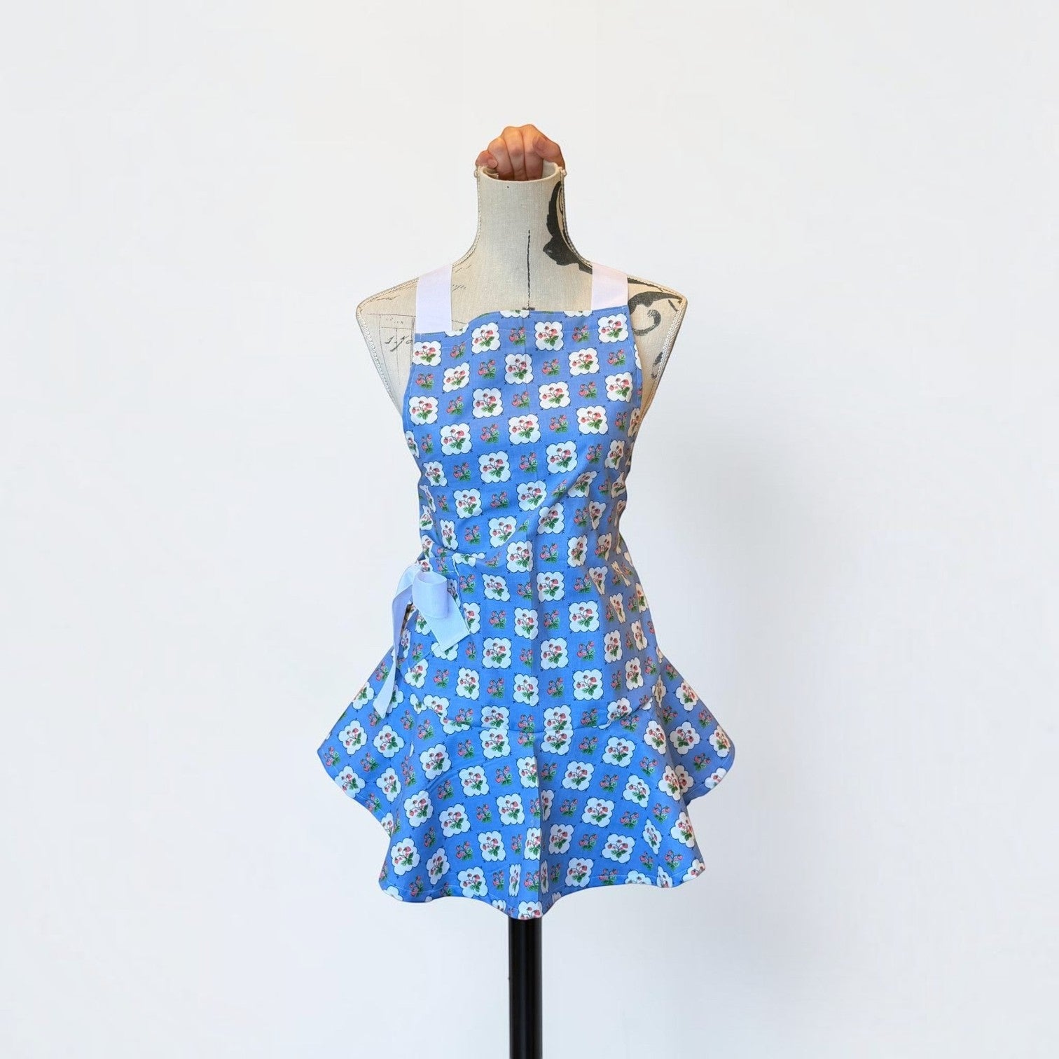 Her Good Eye vintage-inspired blue floral apron with a flared peplum hem, white straps, and adjustable waist tie.