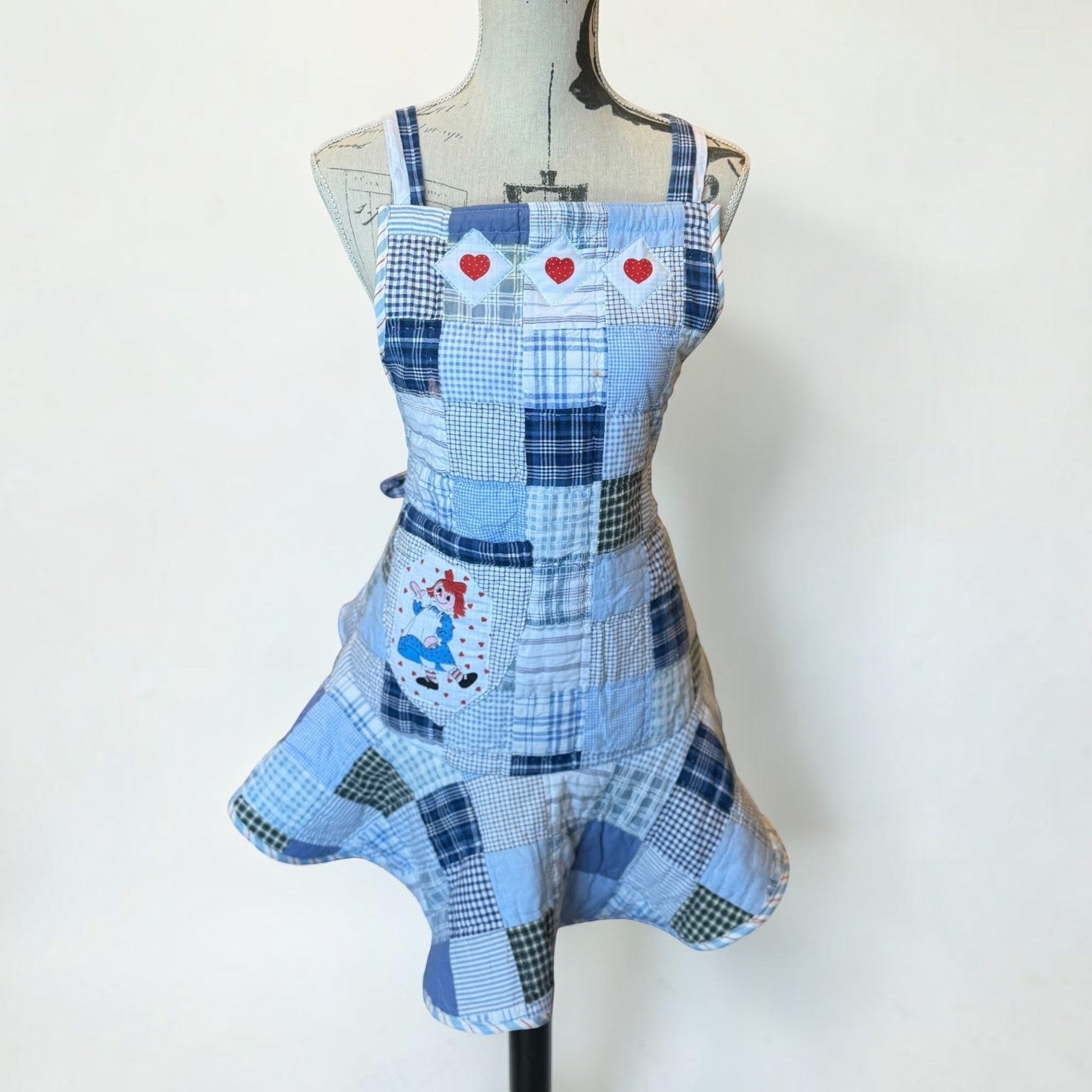 Her Good Eye blue quilted patchwork apron with red heart appliqués and a vintage character patch on a mannequin.