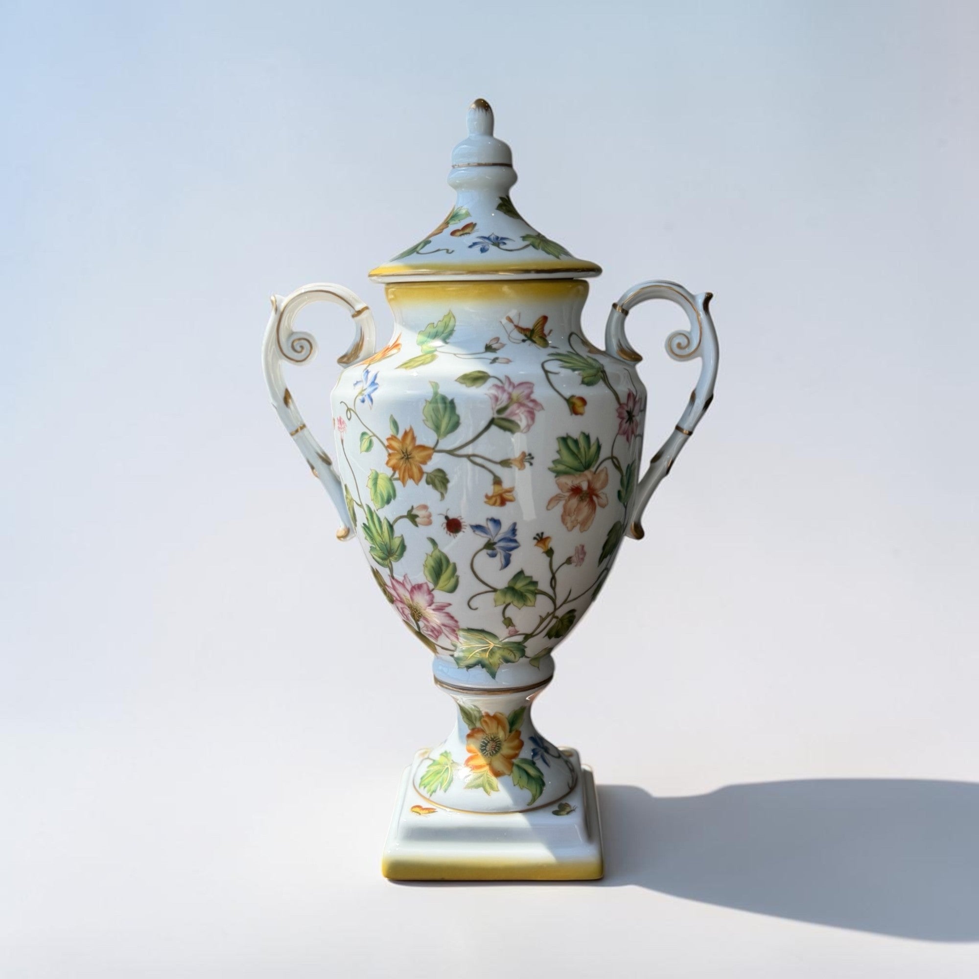 Her Good Eye decorative white ceramic urn with lid, hand-painted with colorful floral motifs and gold trim.