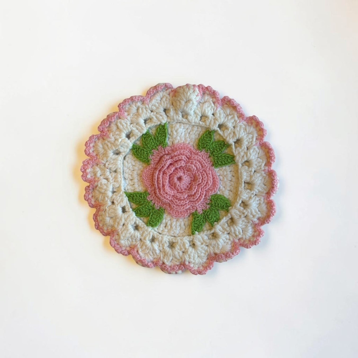 Her Good Eye handmade crochet coaster with a pink rose, green leaves, and cream scalloped edge in cottagecore style.