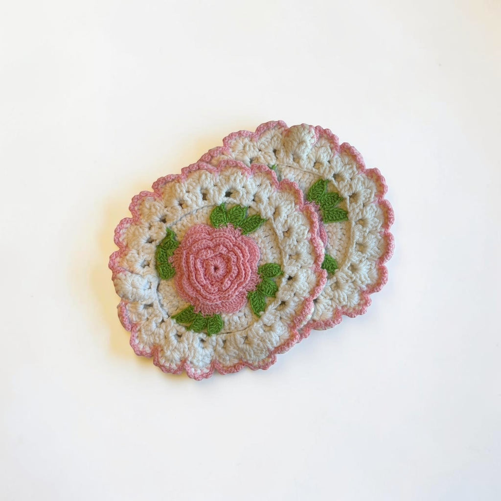 Her Good Eye handmade crochet coaster with a pink rose, green leaves, and scalloped pink trim on a cream base.