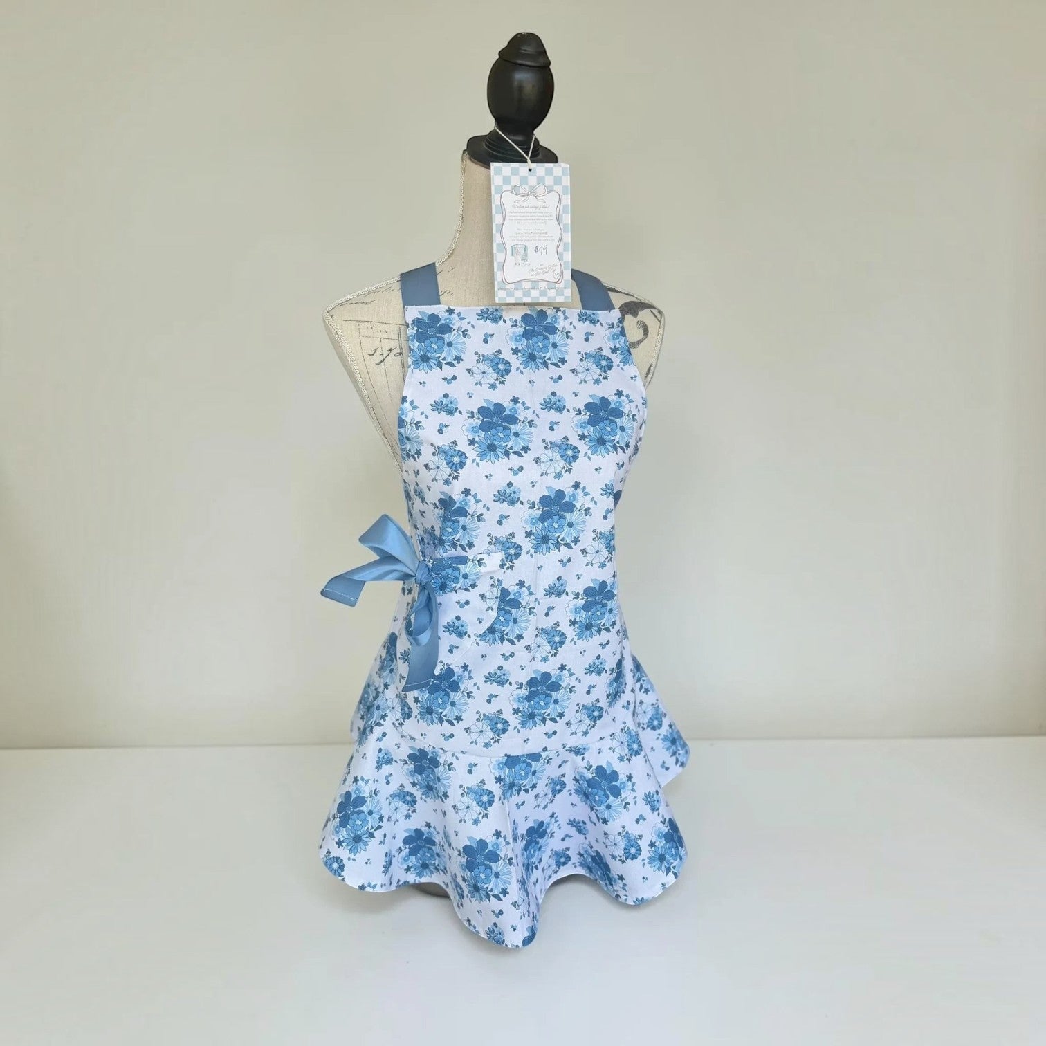 Her Good Eye floral print kitchen apron in blue and white with a ruffled hem and satin ribbon ties on a mannequin.