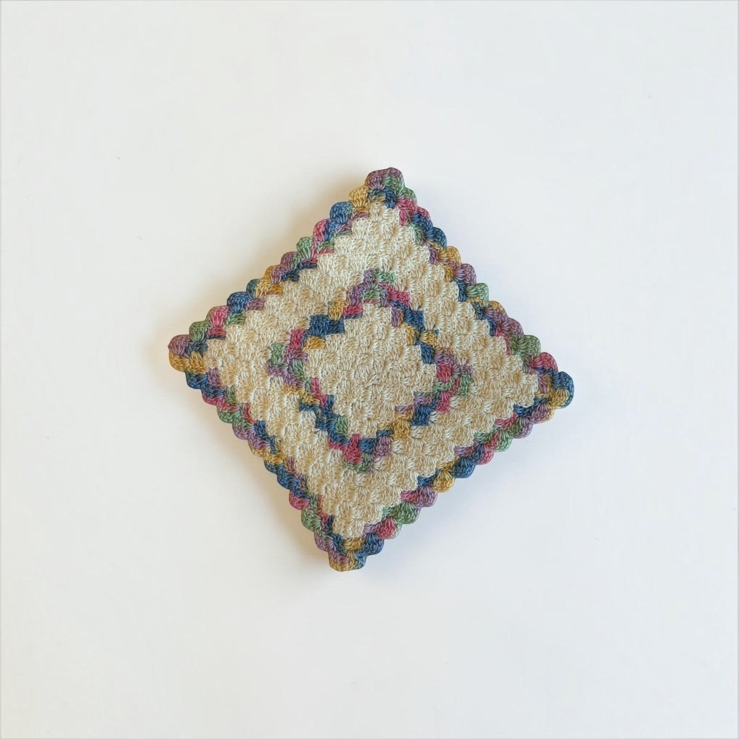 Her Good Eye handmade square crochet drink coaster in cream cotton with colorful blue, pink, green, and gold accents.
