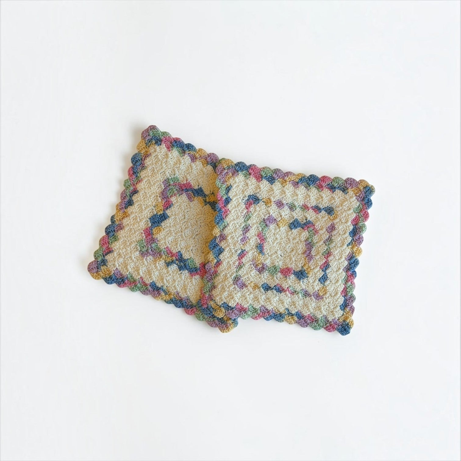 Her Good Eye handmade square crochet drink coaster in cream cotton with multi-colored blue, pink, green, and gold yarn.