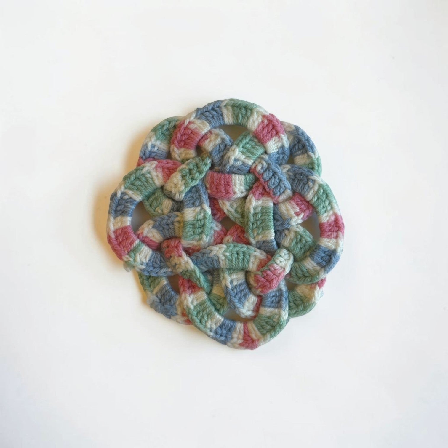 Her Good Eye handmade crochet coaster in variegated sage green, dusty rose, and slate blue with an intricate knot design