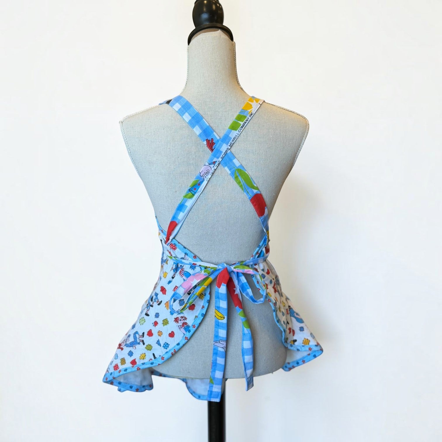 Her Good Eye quilted kitchen apron with cartoon character print, blue gingham straps, and ruffled skirt back view.
