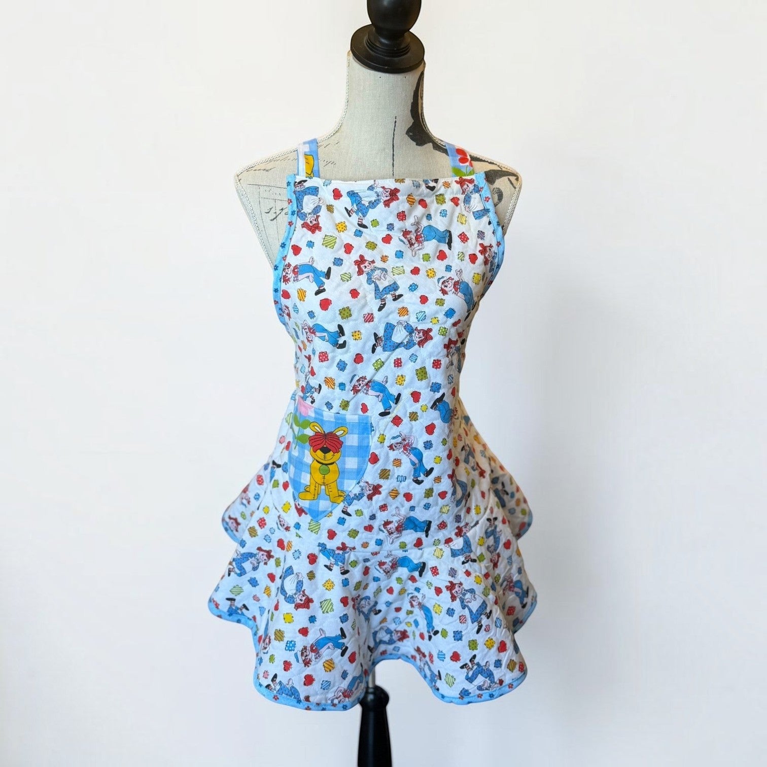 Her Good Eye quilted kitchen apron with multi-colored cartoon character print, blue gingham pocket, and ruffle skirt.