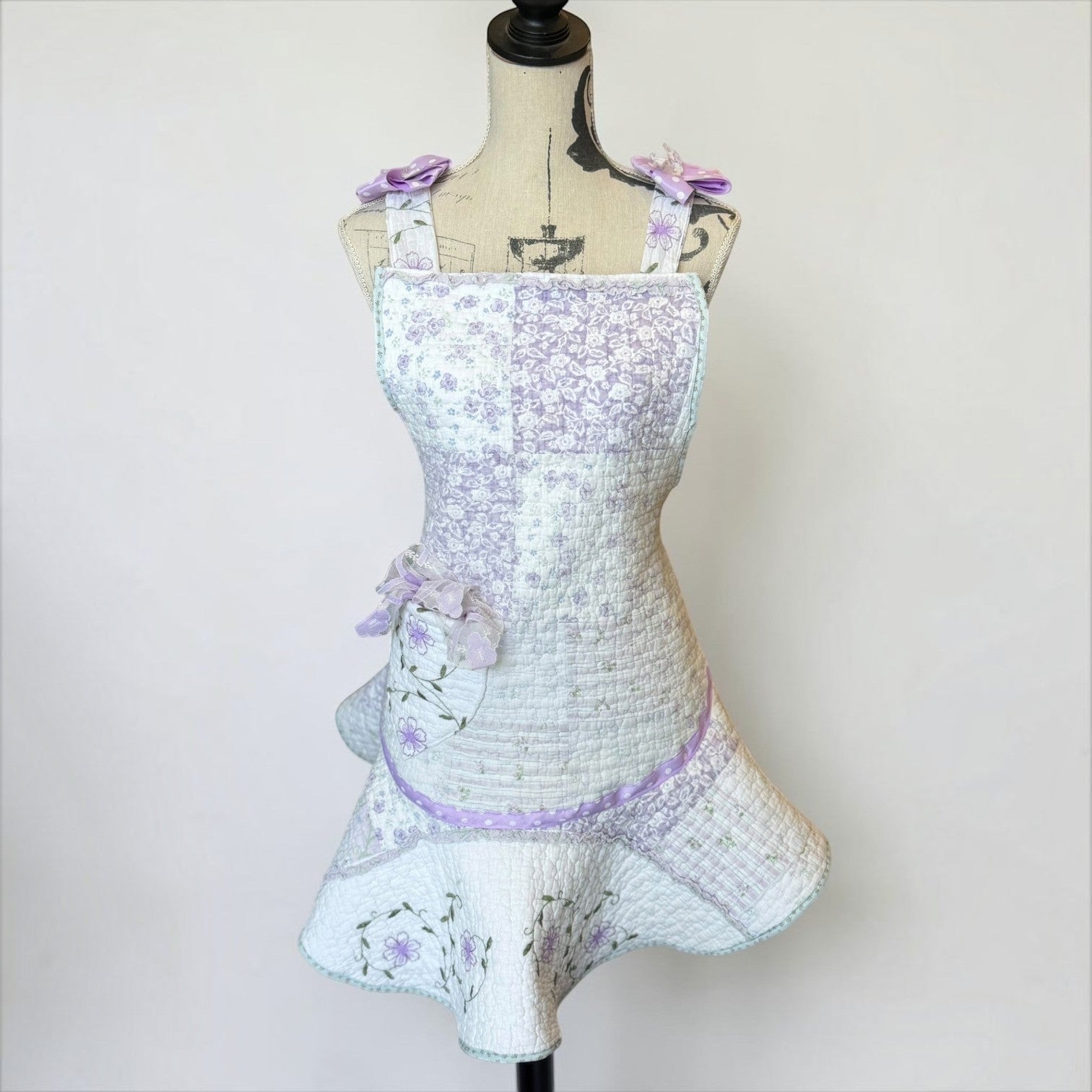 Her Good Eye quilted floral patchwork apron in lavender and white with ribbon bows and a flared hem on a mannequin.