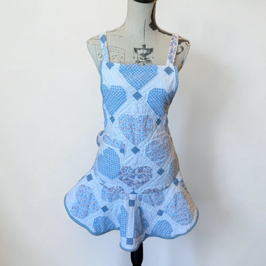 Her Good Eye quilted kitchen apron featuring blue and white heart patchwork, floral patterns, and a ruffled hem.