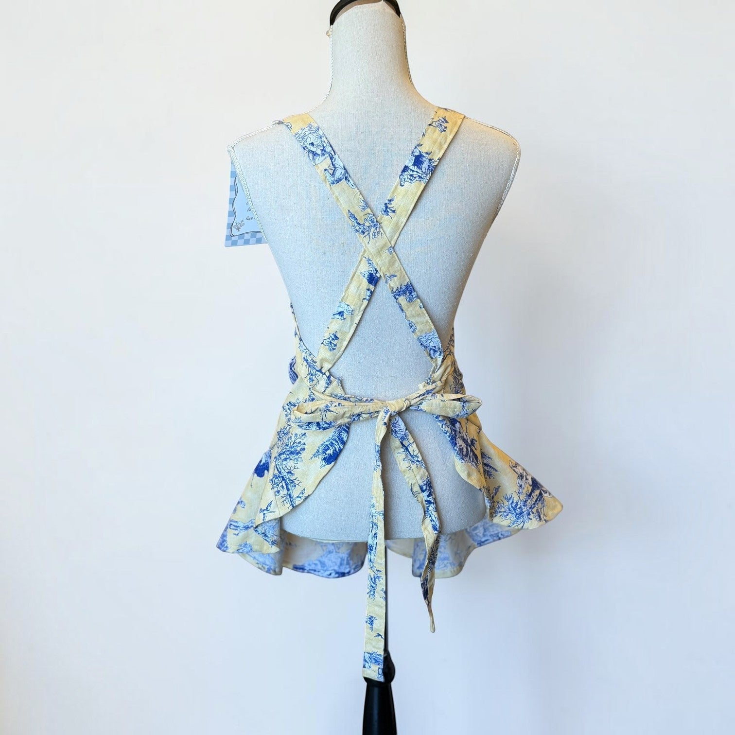 Her Good Eye blue and yellow toile print kitchen apron with a ruffled hem and cross-back tie closure on a mannequin.