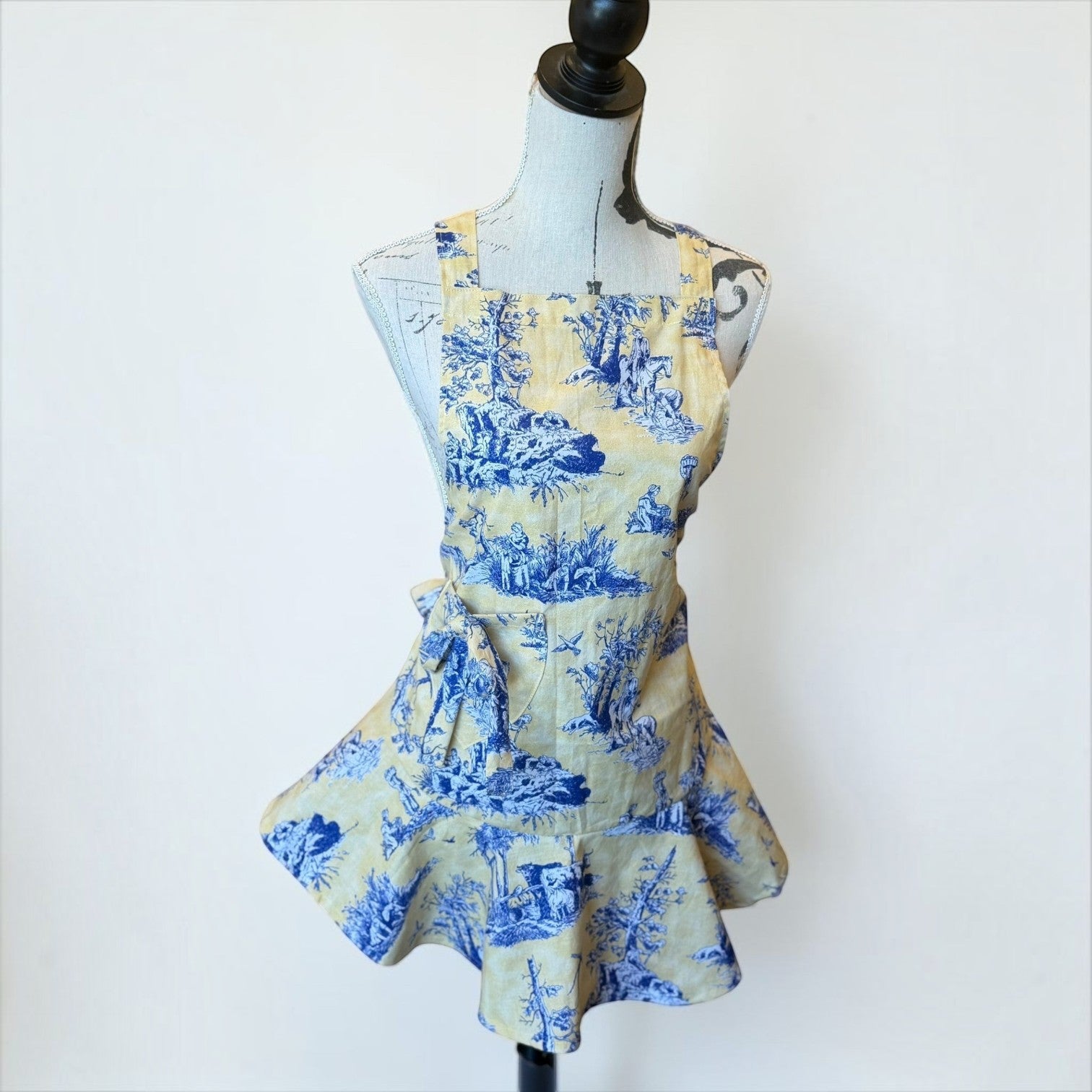Her Good Eye kitchen apron in blue and yellow toile print with a ruffled hem on a dress form mannequin