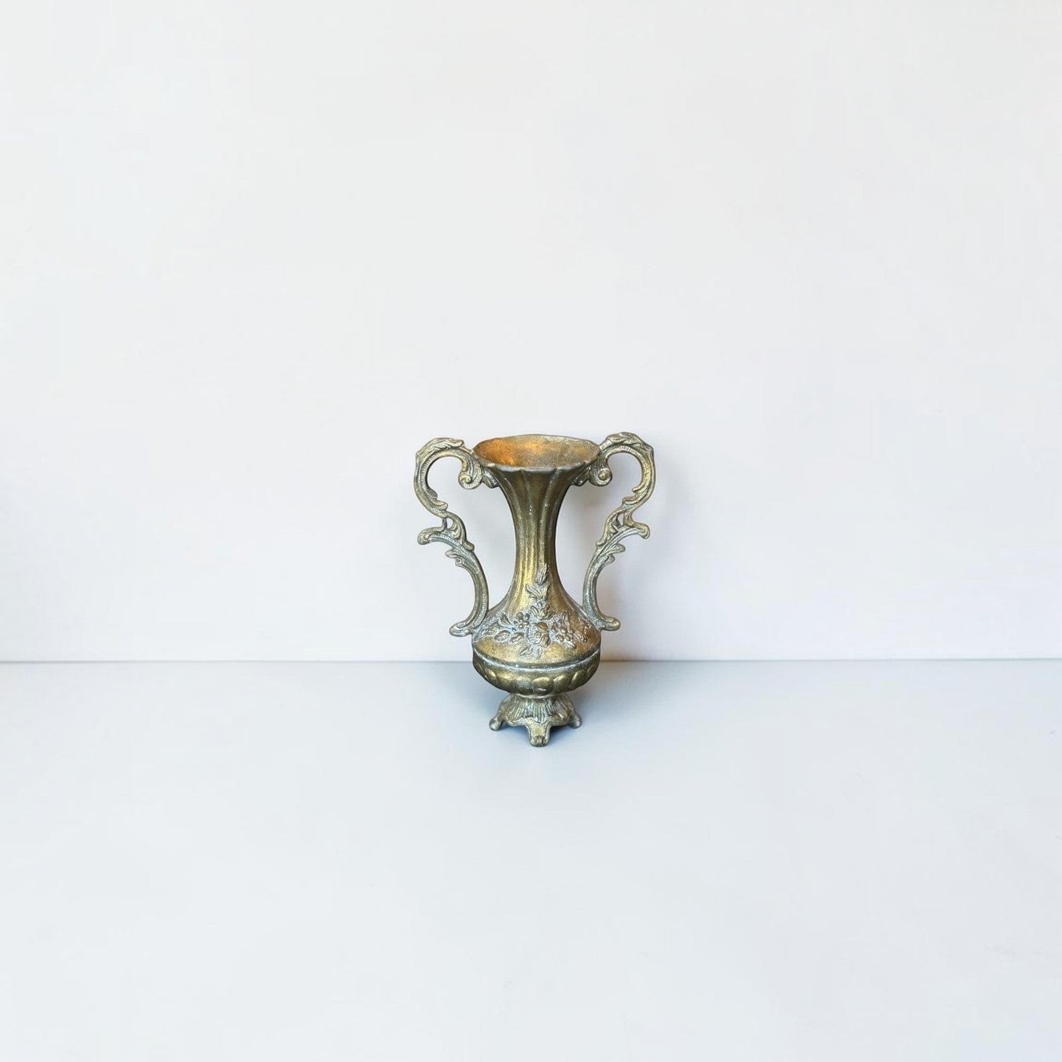 Her Good Eye vintage brass-toned metal urn vase with ornate scroll handles and delicate floral relief detailing.