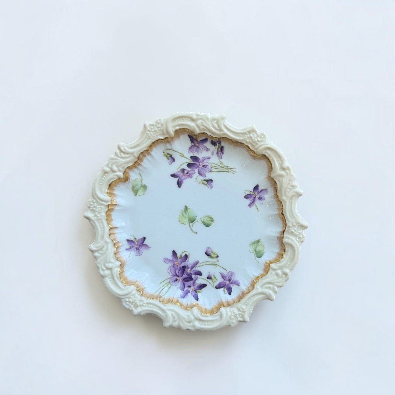 Her Good Eye vintage porcelain plate with hand-painted violet flowers, scalloped relief border, and gold-toned rim.